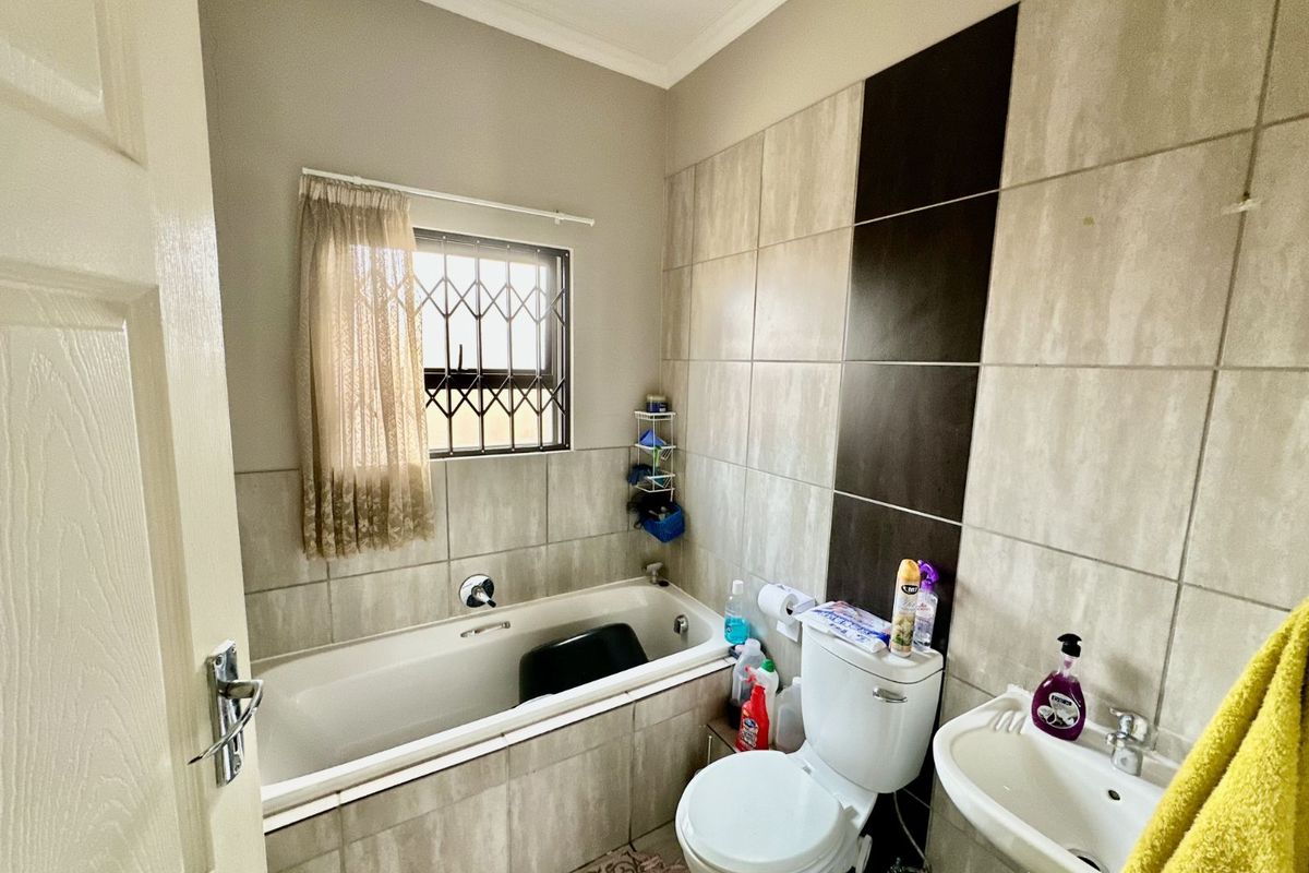 A functional bathroom featuring a built-in bathtub, pedestal basin, and toilet with neutral wall tiling and security bars on the window.