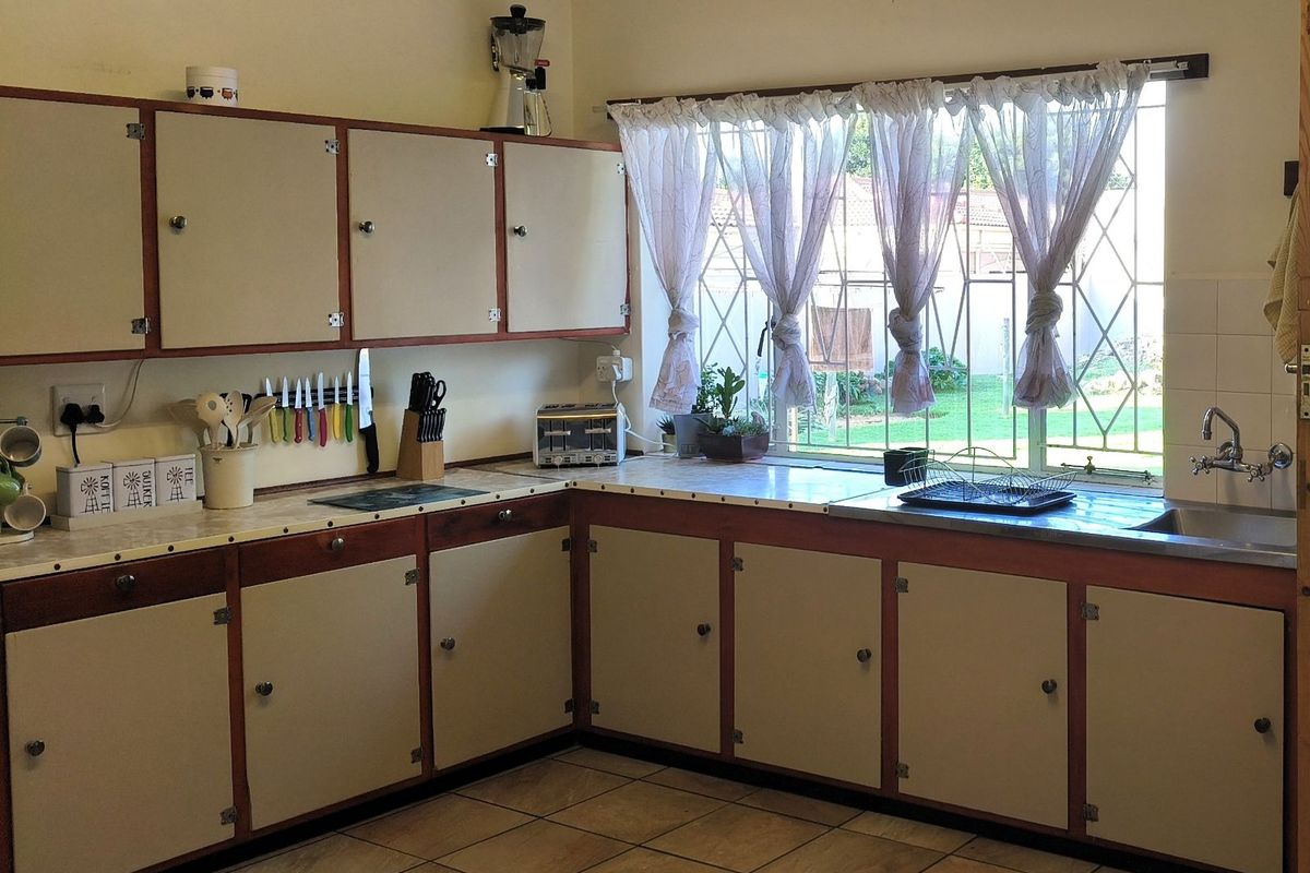 A functional kitchen featuring traditional cabinetry, laminate countertops, a stainless steel sink, and a window with security bars overlooking the garden.