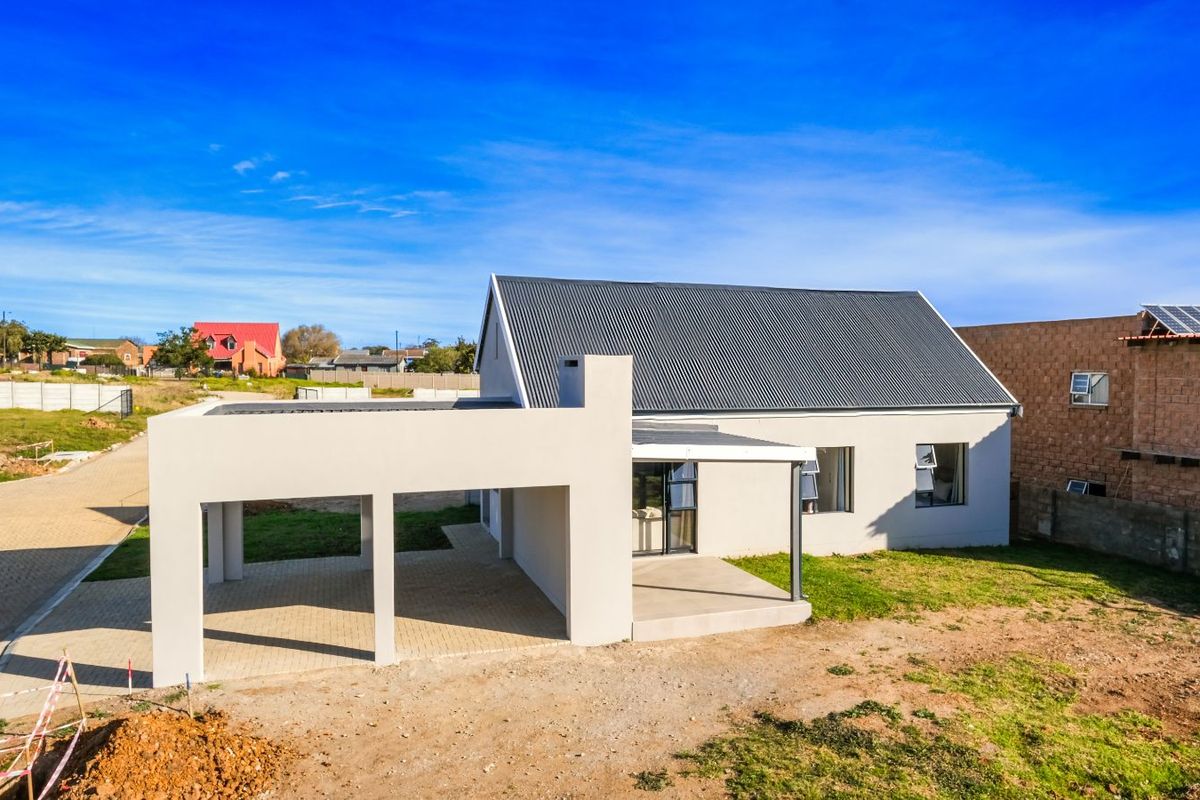 A modern single-story house featuring a corrugated metal roof, a spacious attached carport, and a covered patio area set within a developing residential plot.