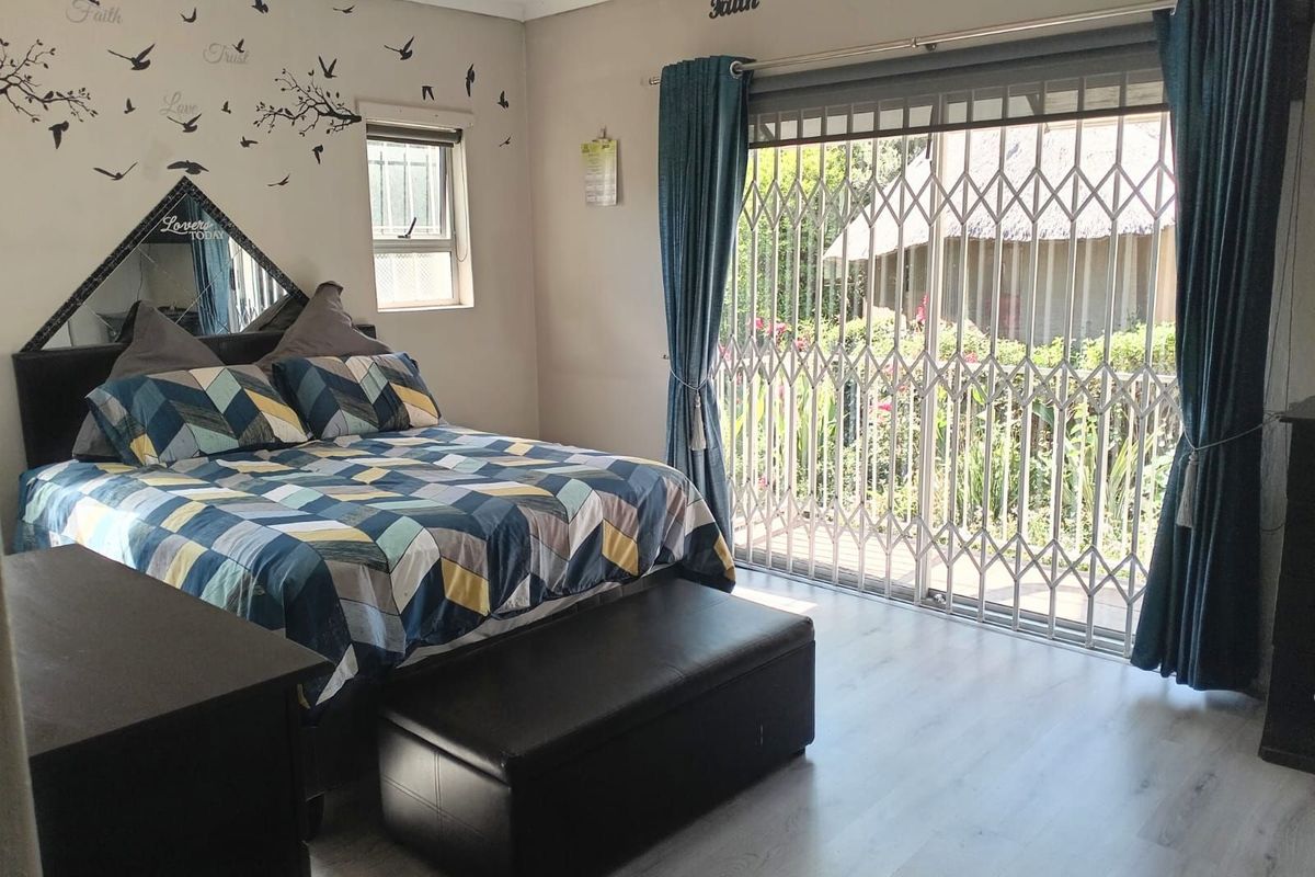 A well-lit bedroom featuring light-toned laminate flooring, a large sliding door with security gate access to the garden, and a small side window.
