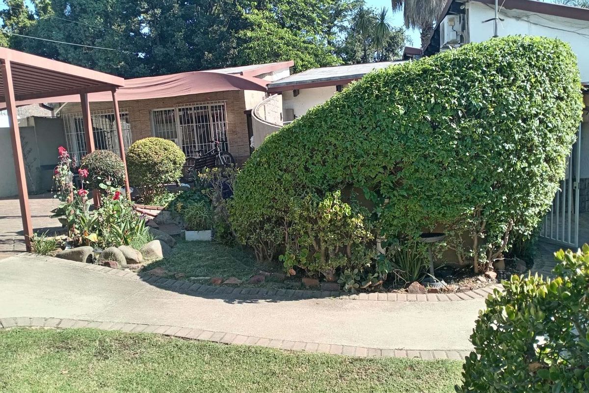 A view of the rear exterior area featuring a covered patio, landscaped garden beds, and a paved walkway leading to the building entrance.