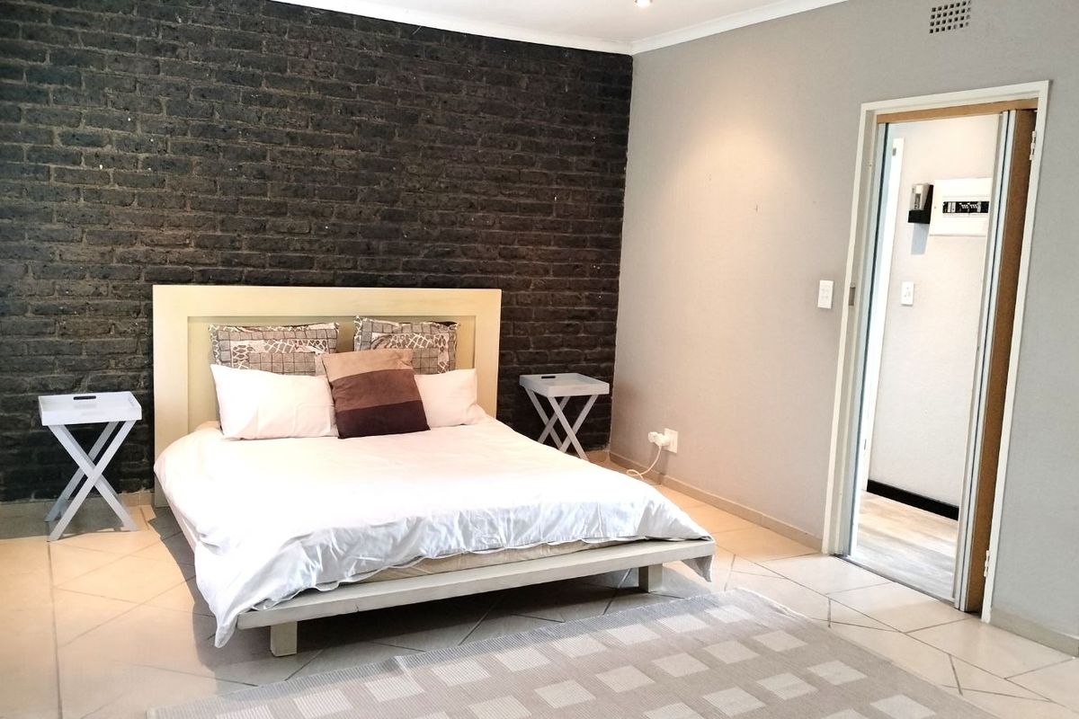 A bedroom featuring a dark exposed brick feature wall, neutral tiled flooring, and an adjoining doorway leading to a secondary space.