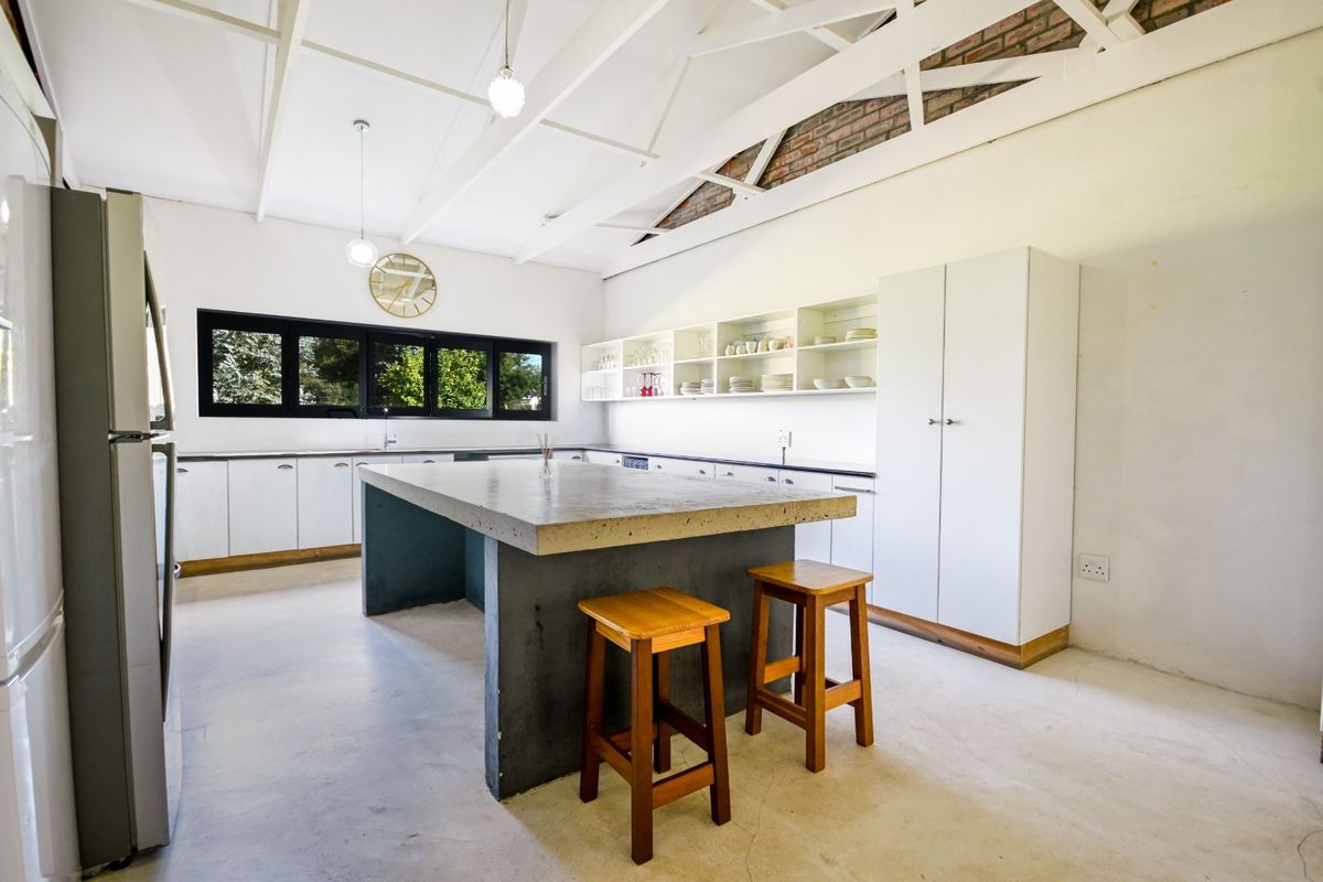 Spacious kitchen featuring a large concrete island, white cabinetry, open shelving, and exposed brickwork under a high vaulted ceiling with exposed trusses.
