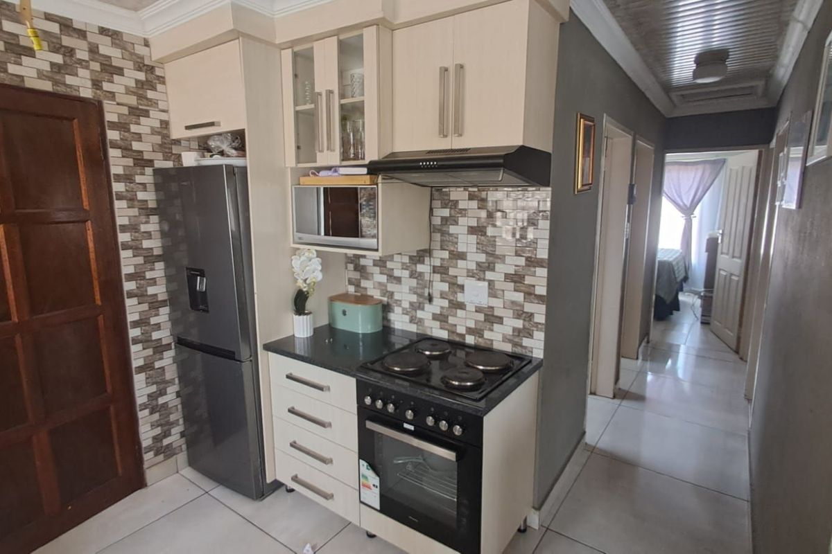 Compact kitchen featuring built-in cabinetry, integrated electric oven and hob, extractor fan, and tiled splashback, leading into a tiled hallway.