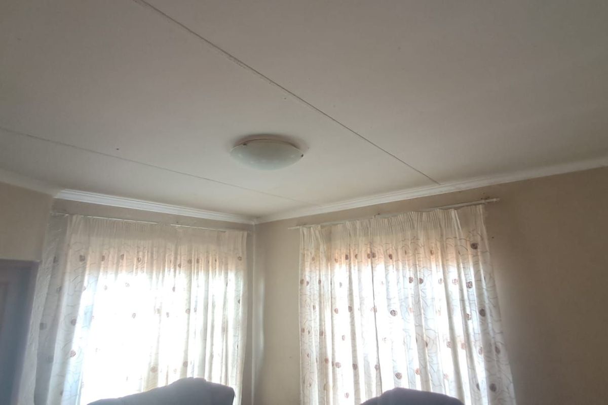A bedroom interior featuring standard ceiling light fixture, curtain rails, and windows with visible wear to the wall and ceiling finishes.