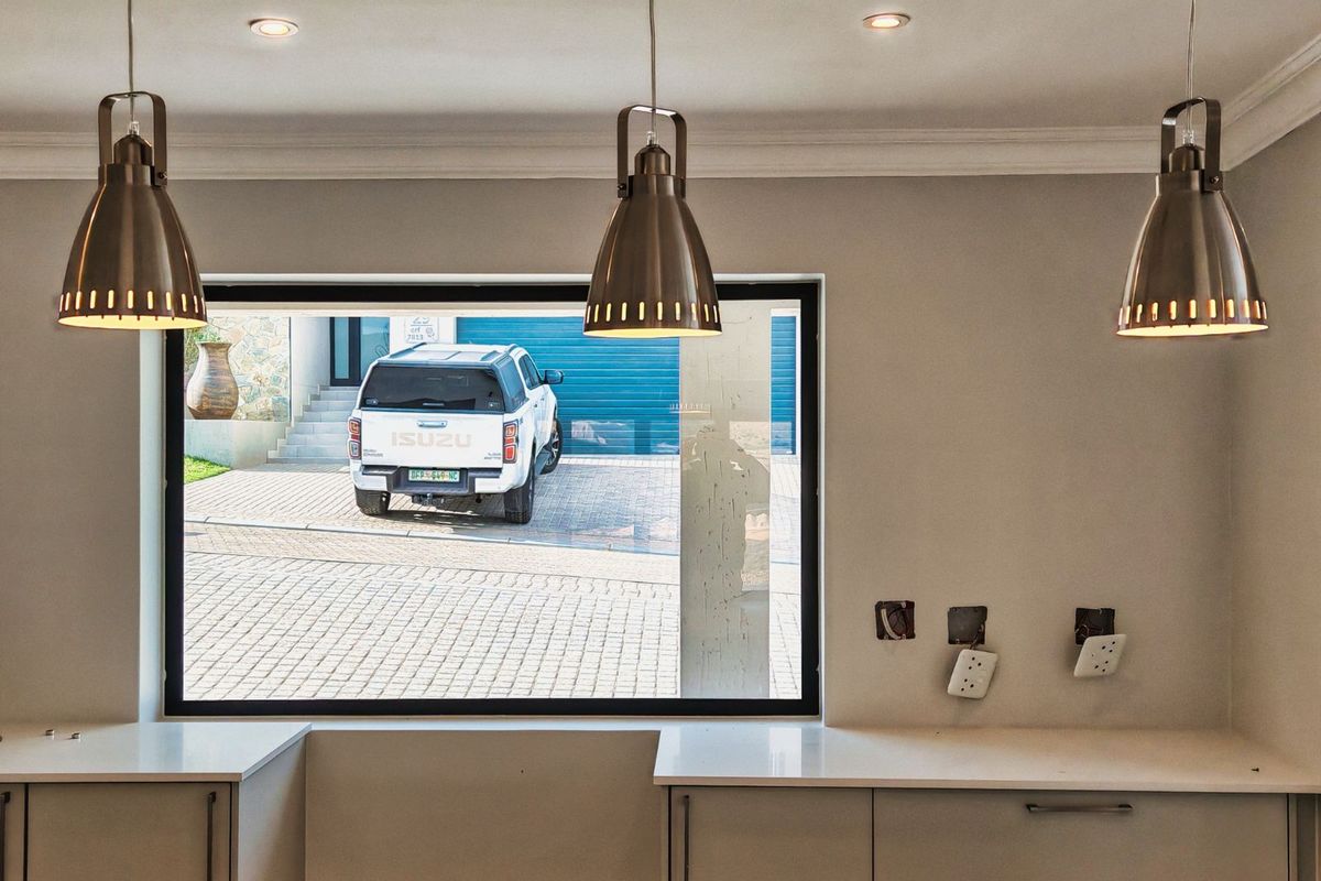 A modern kitchen area featuring sleek cabinetry, stone countertops, and contemporary pendant lighting, with a large window overlooking the driveway.