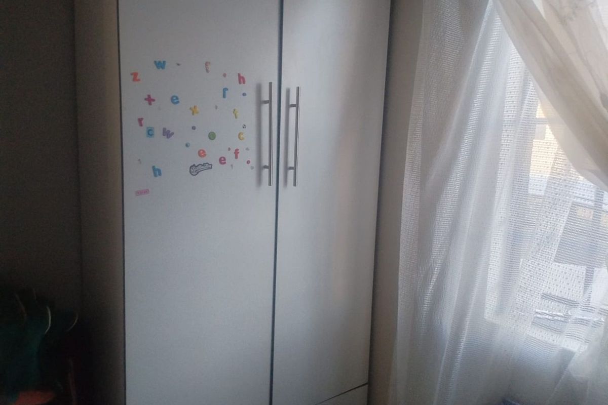 A bedroom featuring a built-in white wardrobe with modern metal handles, positioned next to a window with sheer curtains.
