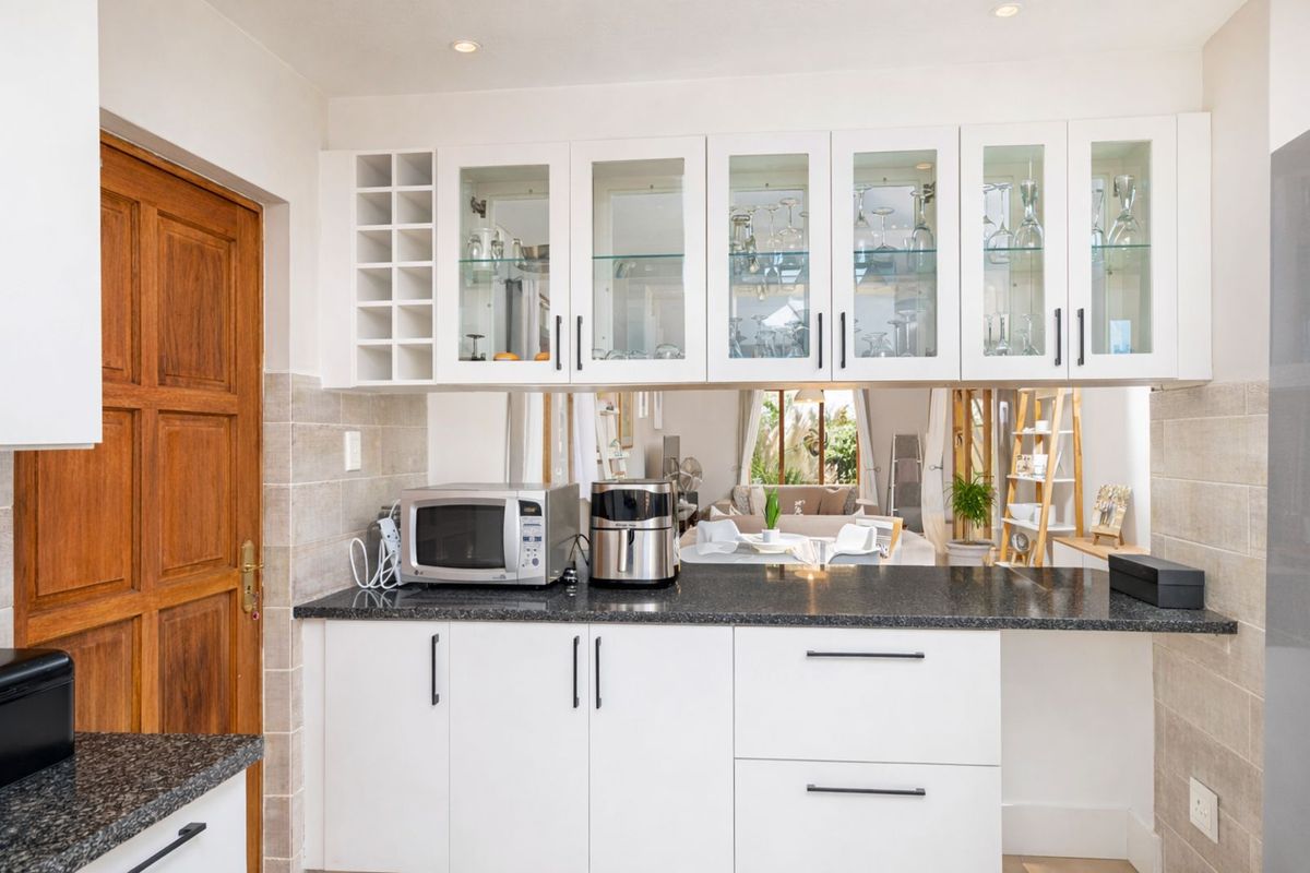 Modern kitchen featuring white cabinetry, glass-front display cupboards, dark granite countertops, and a convenient pass-through window overlooking the living area.
