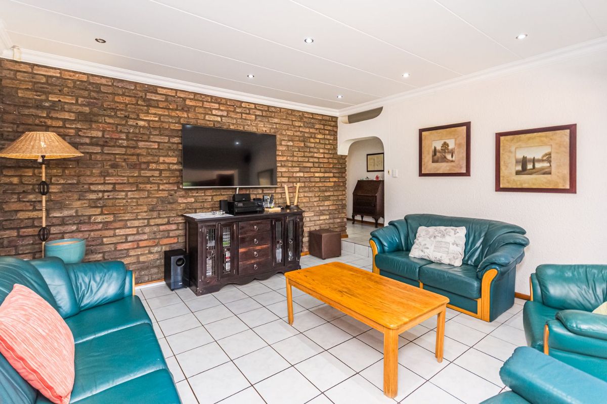 A living room featuring a prominent exposed brick feature wall, tiled flooring, and recessed ceiling downlights, leading into an adjacent hallway.