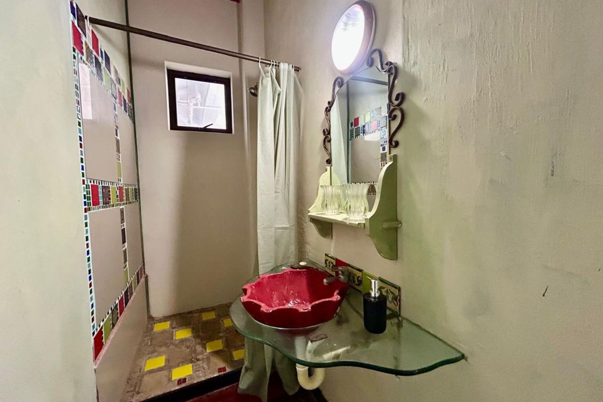 A compact bathroom featuring a unique red vessel sink, glass vanity shelf, and mosaic-tiled shower area with a curtain enclosure.