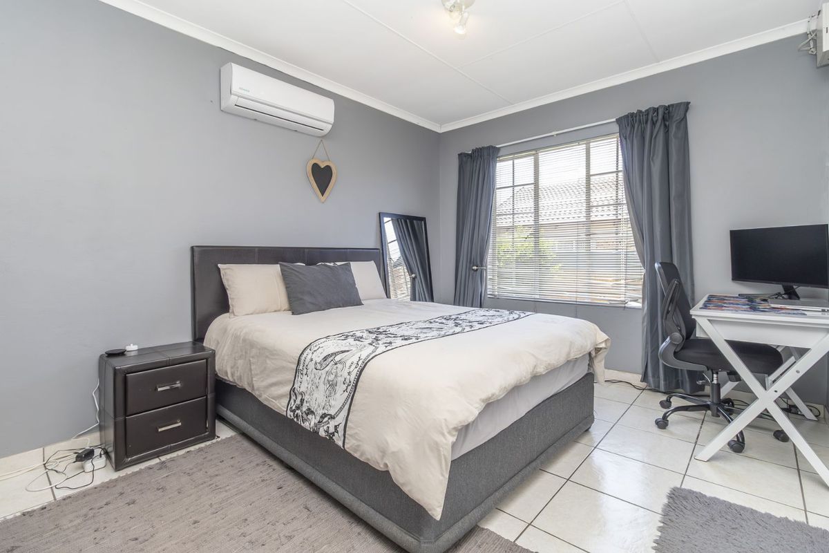 A well-lit bedroom featuring tiled flooring, a wall-mounted air conditioning unit, and large windows with blinds, providing a functional and comfortable living space.