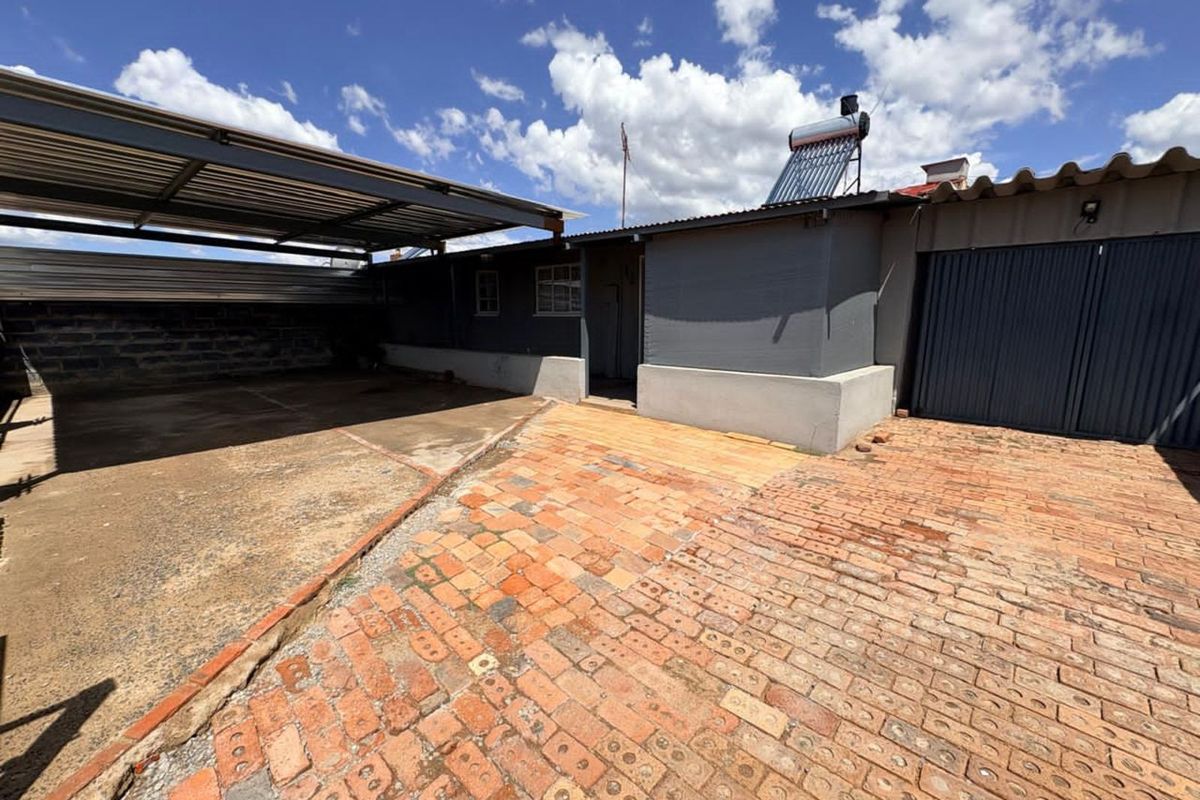 A rear exterior view featuring a covered carport area, brick paved courtyard, and a solar water heating system installed on the roof.