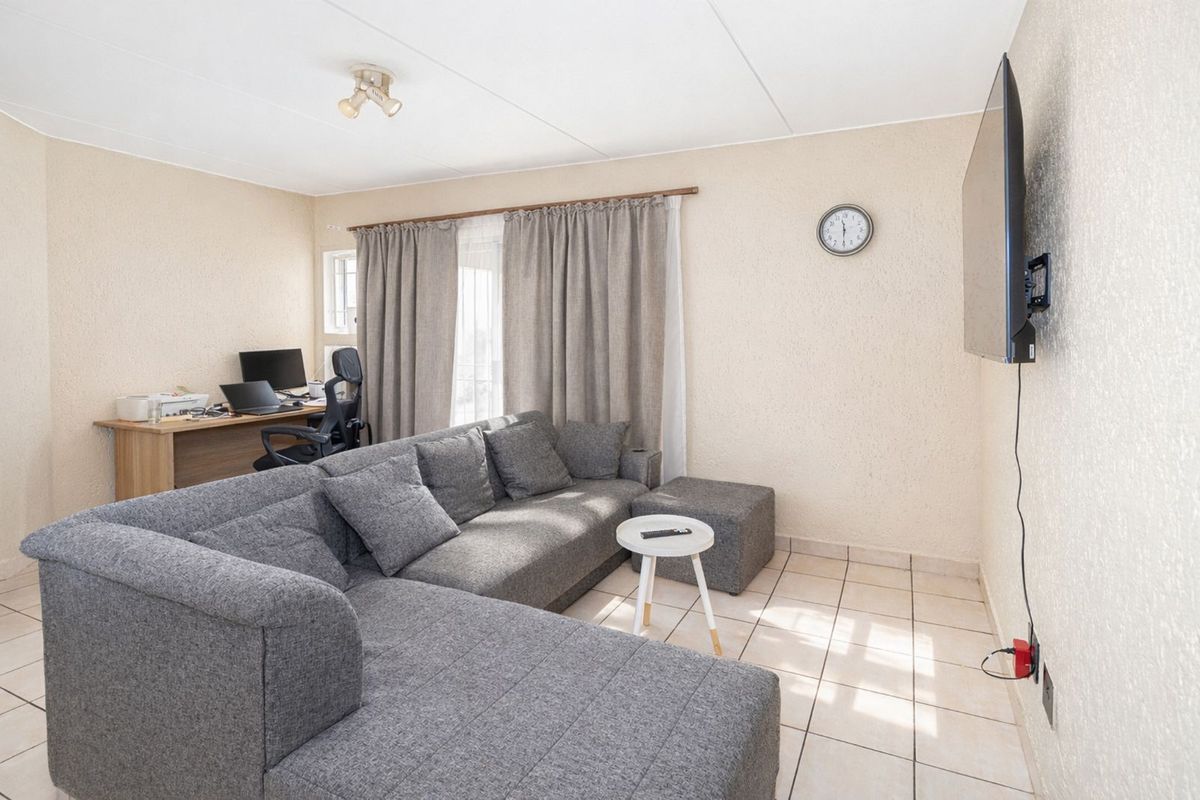 A functional living room featuring tiled flooring, textured walls, and a wall-mounted television, with an integrated workspace area near the window.