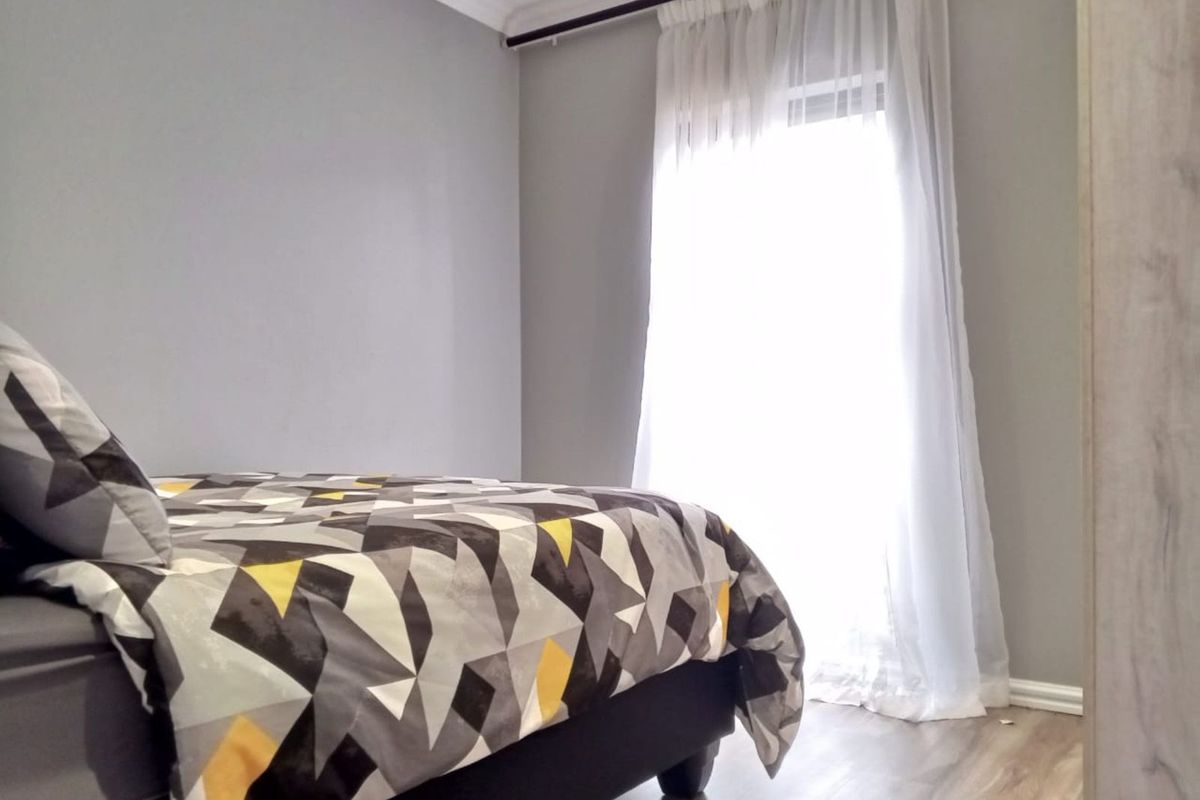 A bright bedroom featuring modern wood-look flooring, neutral grey walls, and a large window with sheer curtains allowing for ample natural light.