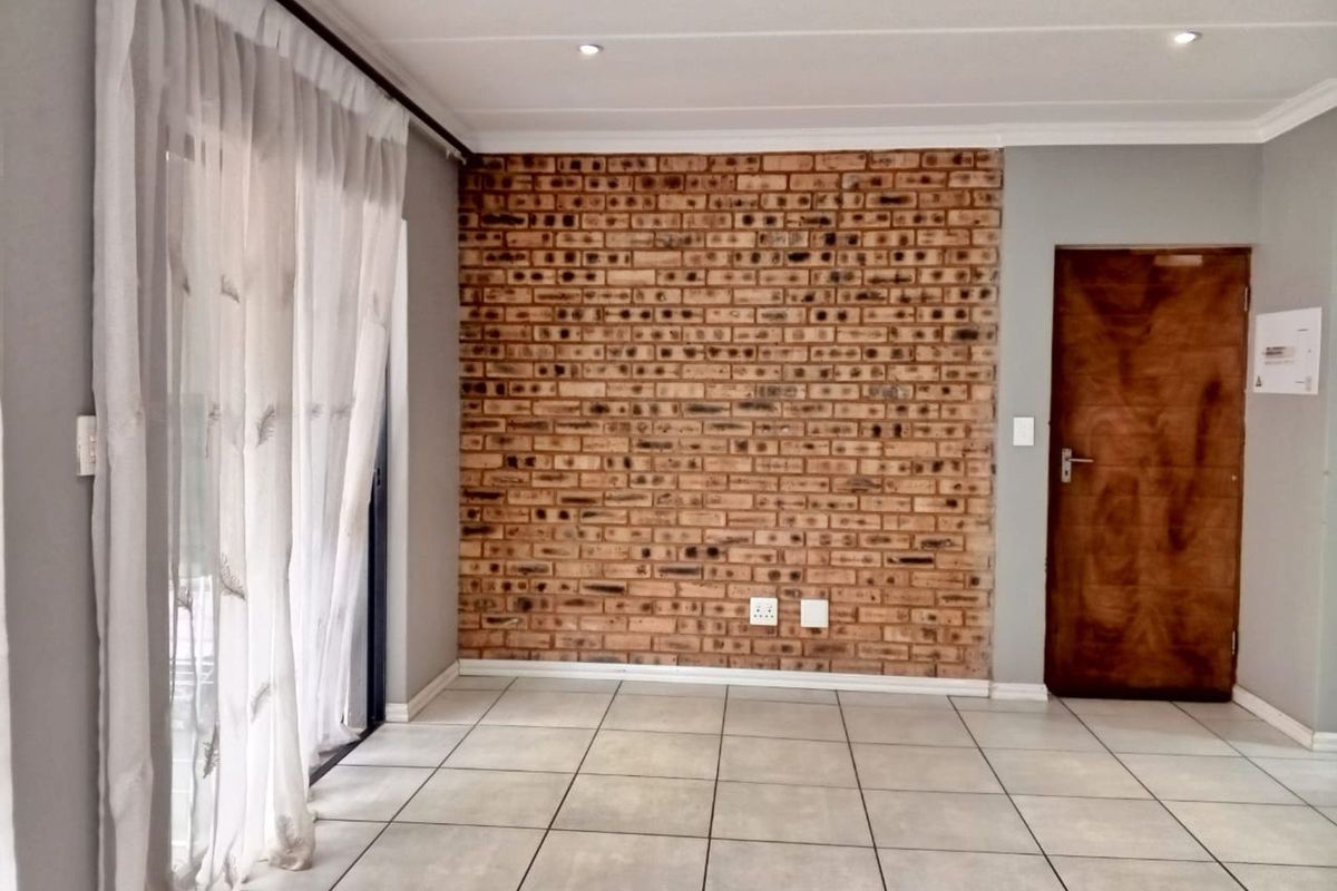 A bright living area featuring a prominent exposed brick feature wall, neutral floor tiles, recessed ceiling lighting, and sliding glass doors leading outdoors.
