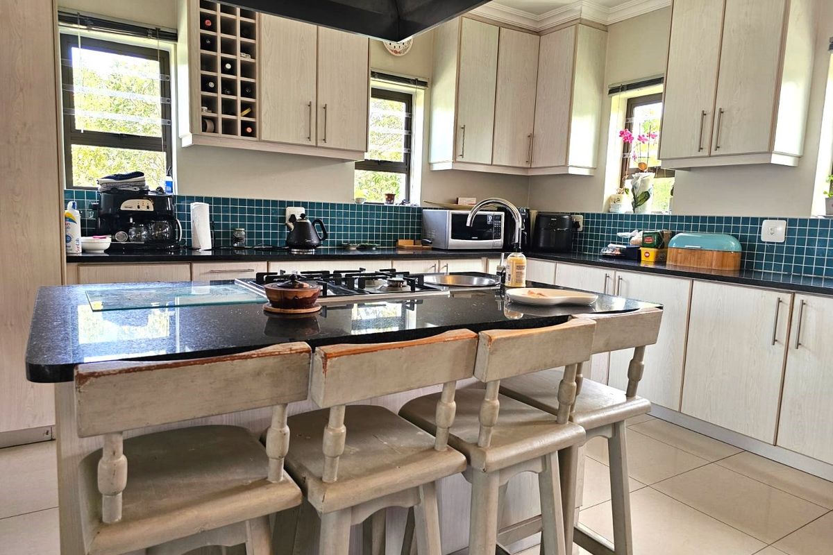 A functional kitchen featuring light wood-grain cabinetry, a central island with a dark stone countertop, a gas hob, and a vibrant teal tiled backsplash.