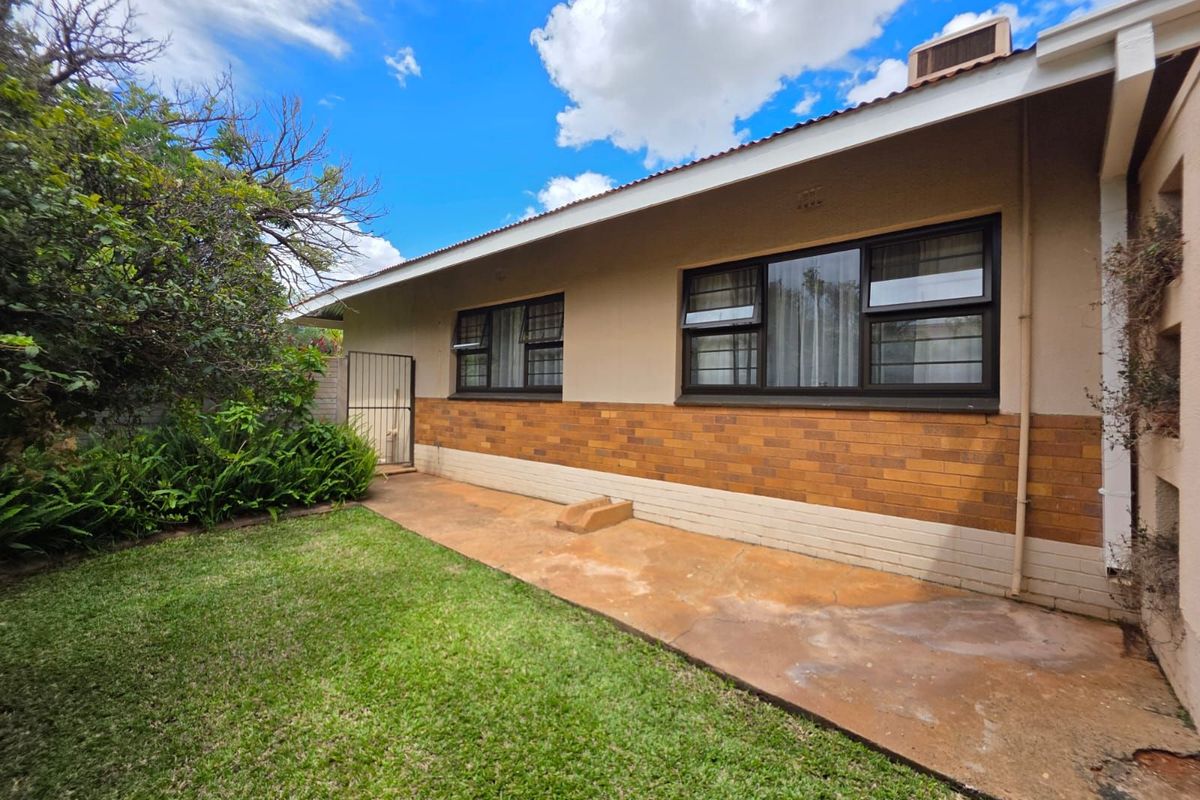 A view of the rear exterior of the house featuring a concrete patio, brick facade, and metal-framed windows overlooking a garden area.