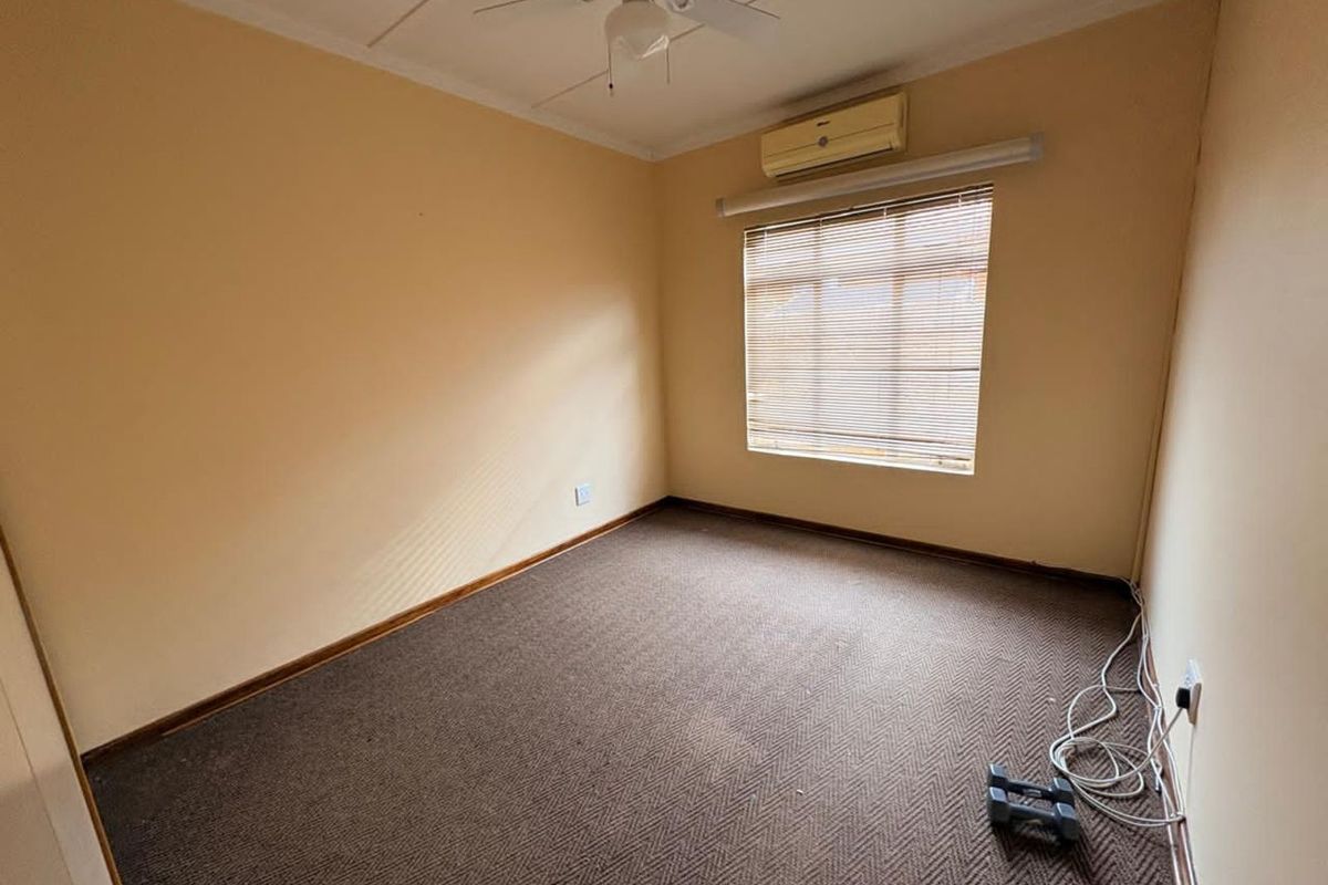 A simple, empty bedroom featuring neutral walls, carpeted flooring, a ceiling fan, and a wall-mounted air conditioning unit.