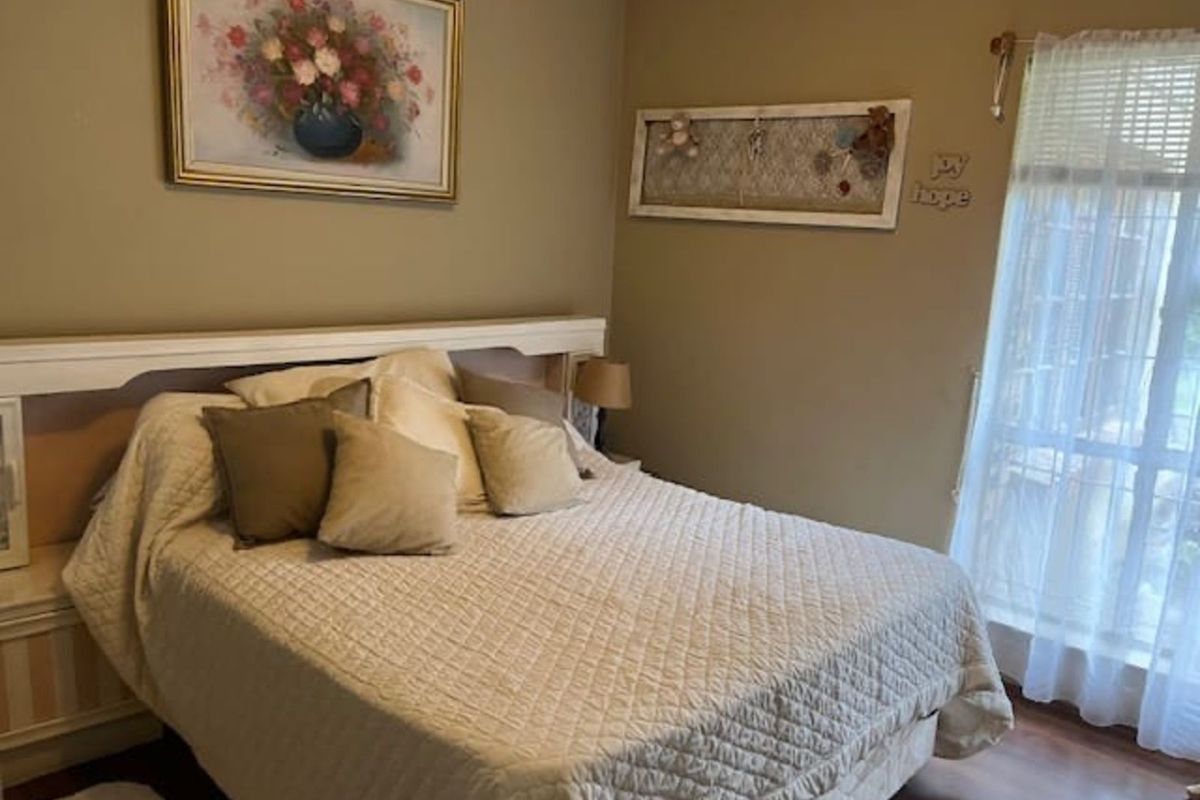 A cozy bedroom featuring a built-in headboard, laminate flooring, and a large window with sheer curtains allowing natural light.