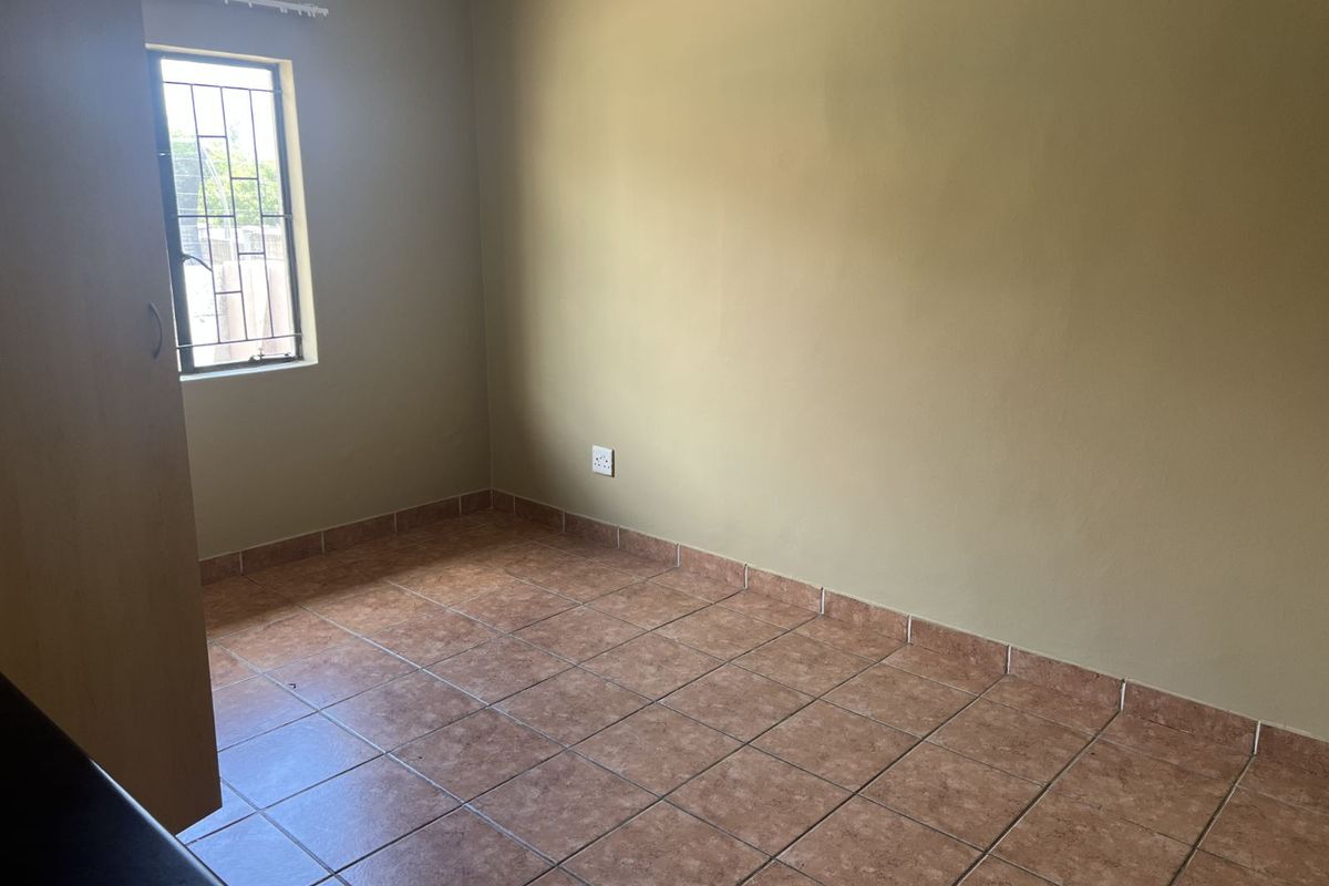 A simple, empty bedroom featuring tiled flooring, a built-in cupboard, and a window with security bars.