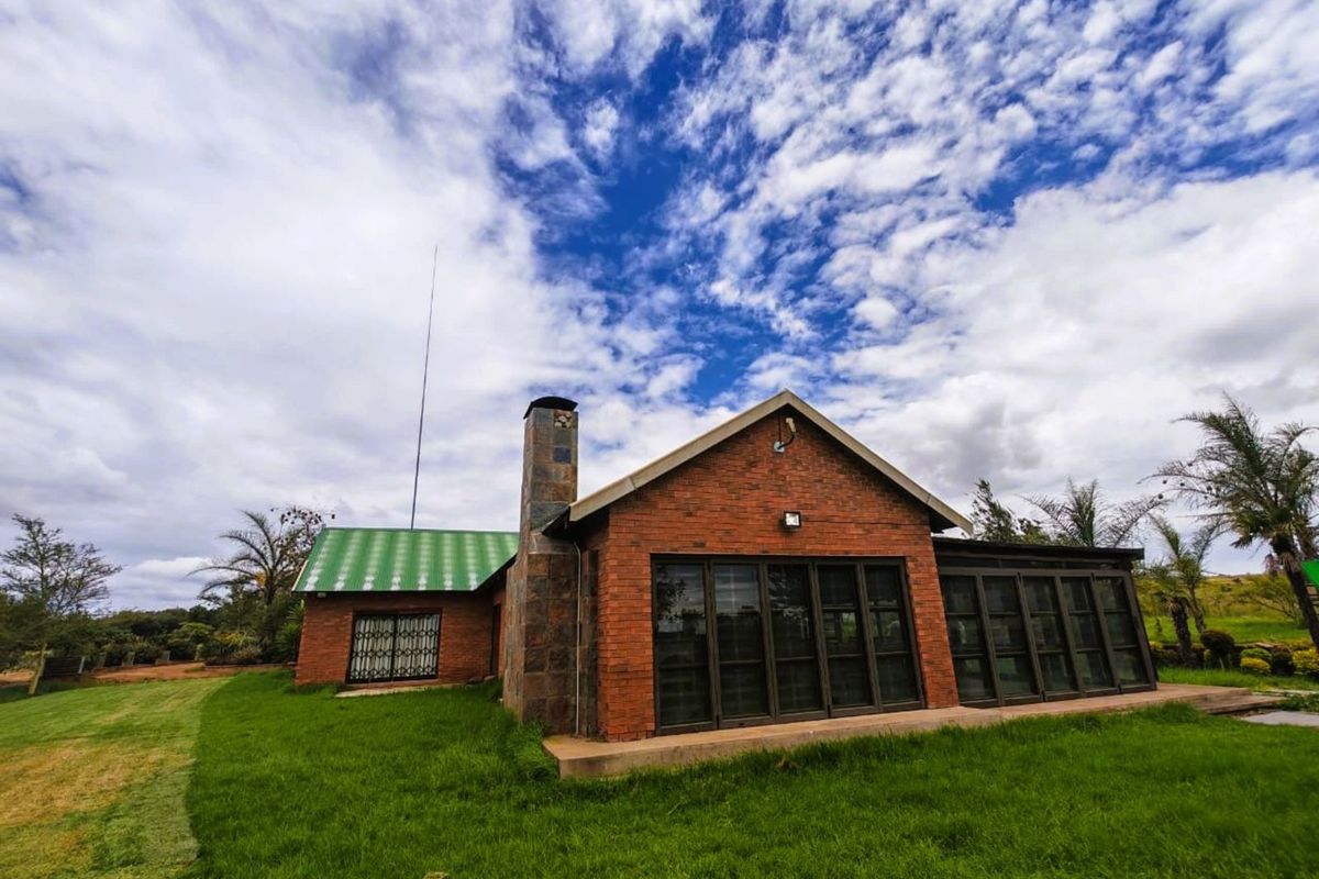A charming brick farmhouse exterior featuring a prominent stone-clad chimney, green metal roof, and expansive glass sliding doors opening onto the surrounding lawn.