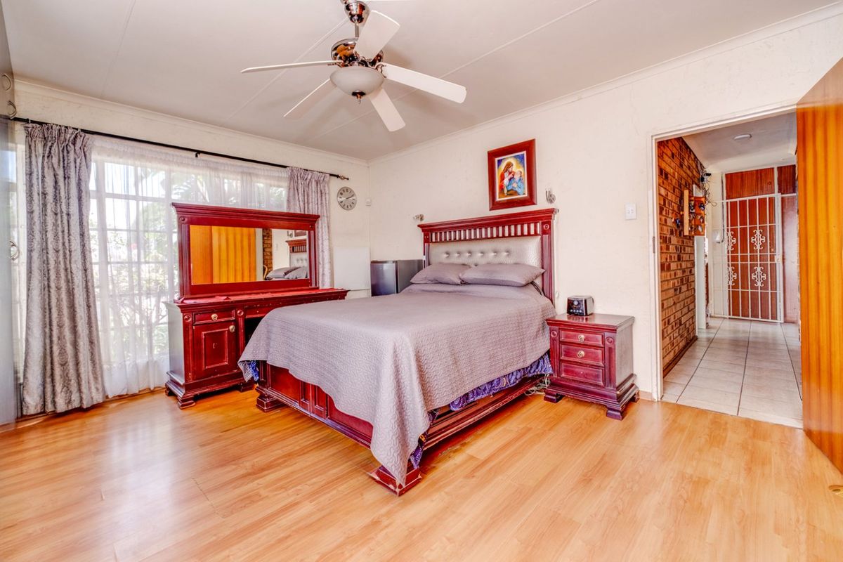 A spacious bedroom featuring laminate flooring, a ceiling fan, and large windows with natural light, leading into a tiled hallway.