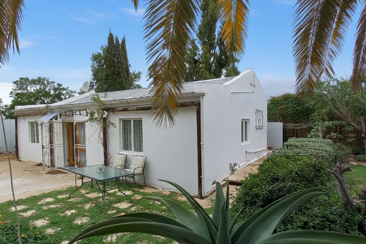 A view of the guest house exterior featuring a white facade, metal security gates, and a surrounding garden area with established landscaping.