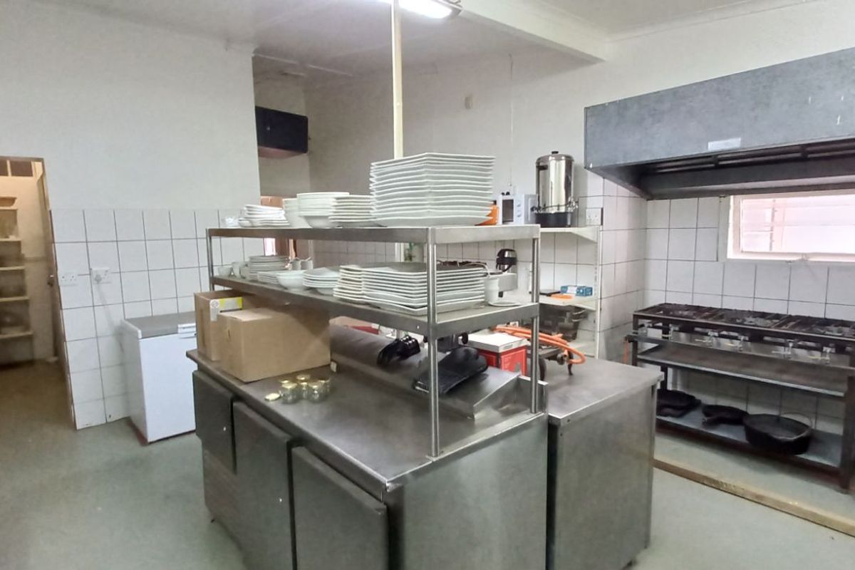Commercial-style kitchen featuring stainless steel prep counters, industrial gas stove, large extractor hood, and tiled walls with an adjacent pantry area.
