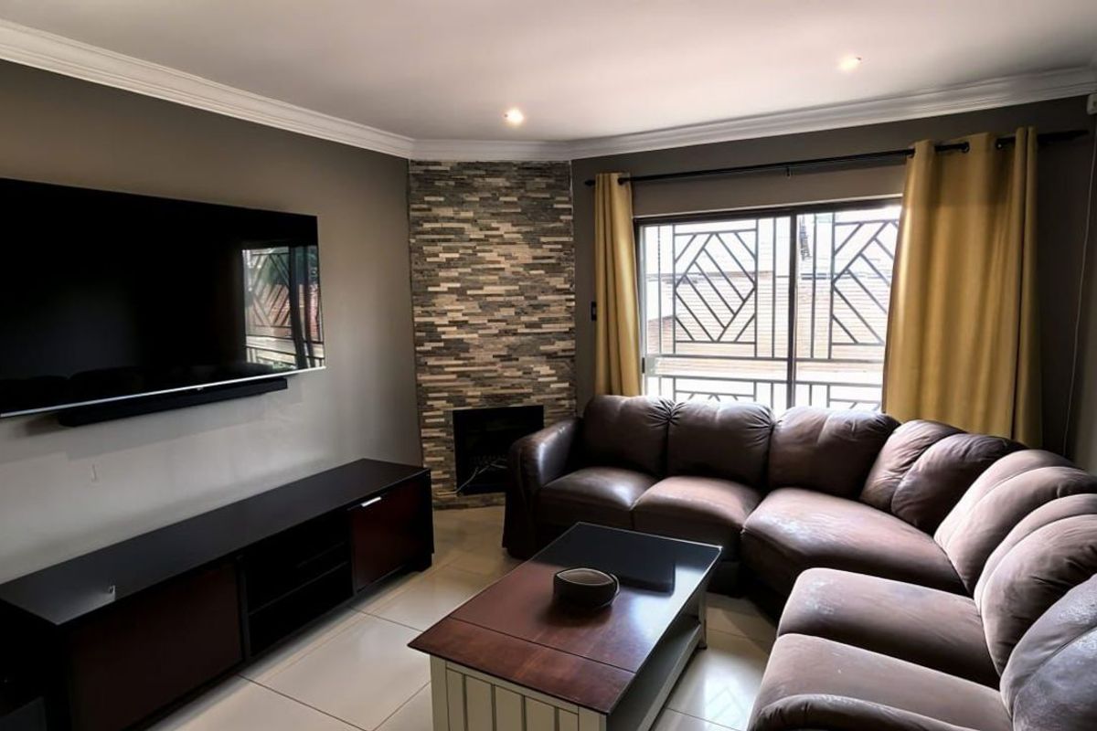 A comfortable living room featuring a corner fireplace with stone cladding, tiled flooring, and large windows with security bars.