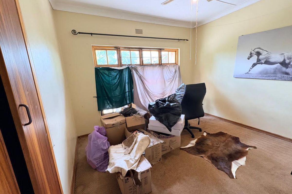 A bedroom featuring built-in cupboards, a ceiling fan, and a window with curtain rod, currently used as a storage space.