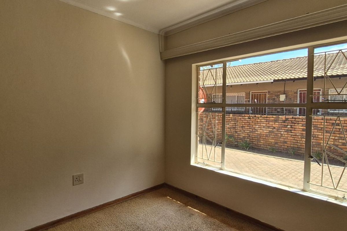 A bright, empty bedroom featuring neutral walls, carpeted flooring, and a large window fitted with security bars overlooking the exterior area.