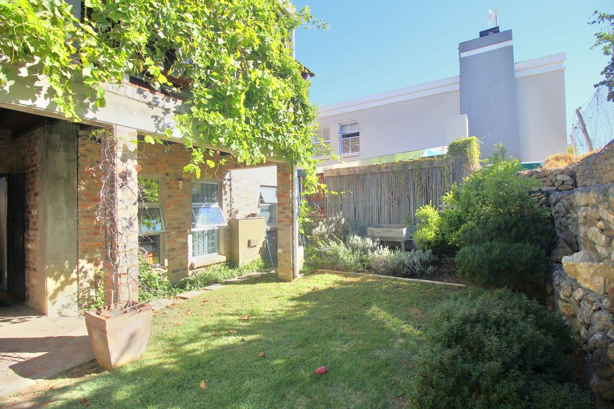 A private rear garden featuring a covered patio area with brick pillars, established greenery, and a stone retaining wall.