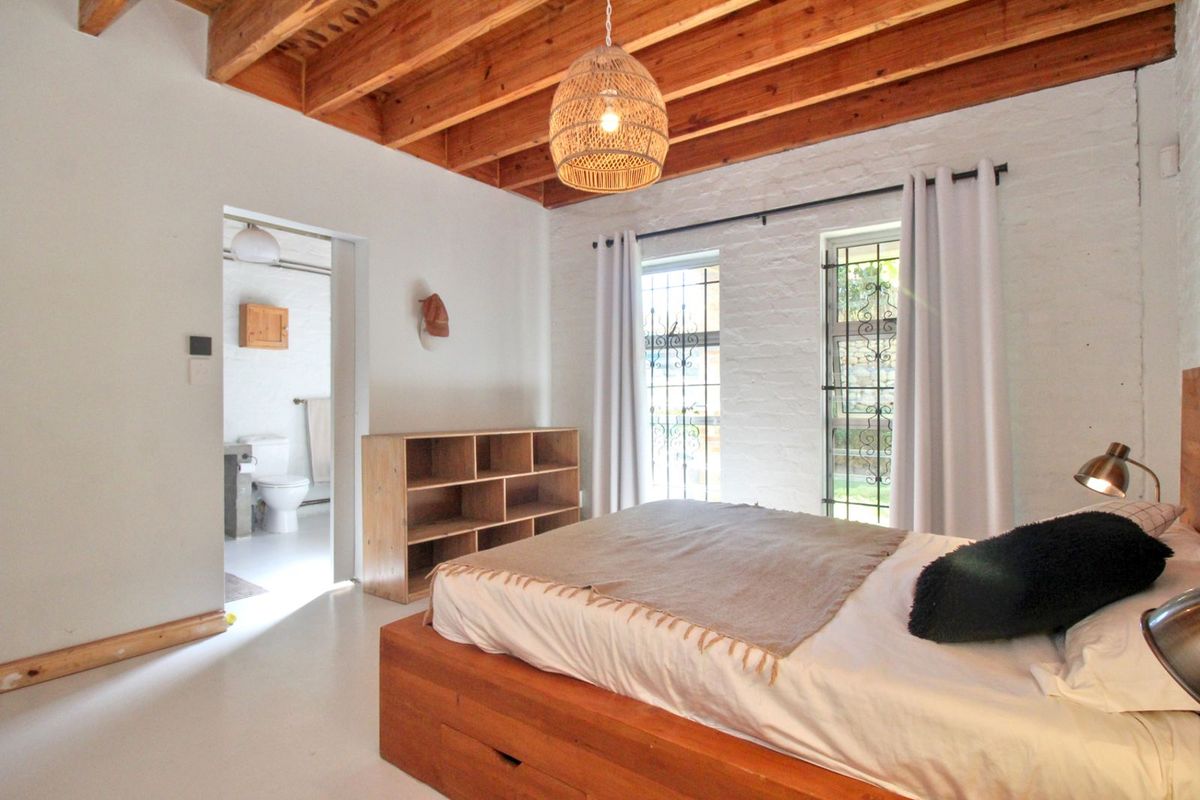 A bright bedroom featuring exposed timber ceiling beams, painted brick walls, and an en-suite bathroom with security bars on the windows.