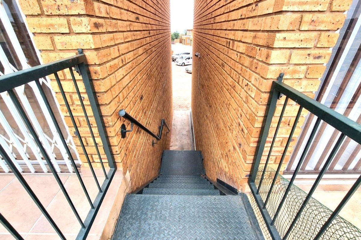 An exterior view of a metal staircase leading down between two brick walls, featuring metal railings and a view towards the parking area.