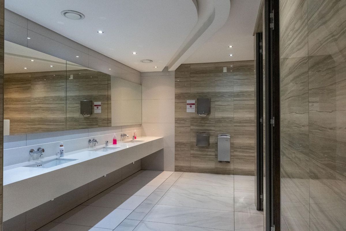 Modern office restroom featuring a long vanity with dual integrated sinks, large wall-mounted mirrors, recessed ceiling lighting, and high-quality tiled wall and floor finishes.