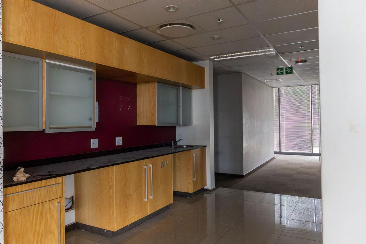 A functional office kitchenette featuring light wood cabinetry, dark countertops, a stainless steel sink, and a suspended ceiling with integrated lighting.
