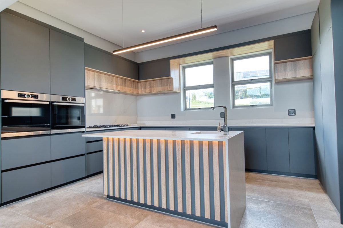 Modern kitchen featuring sleek grey cabinetry, a central island with timber slat detailing, integrated double ovens, and stone countertops.