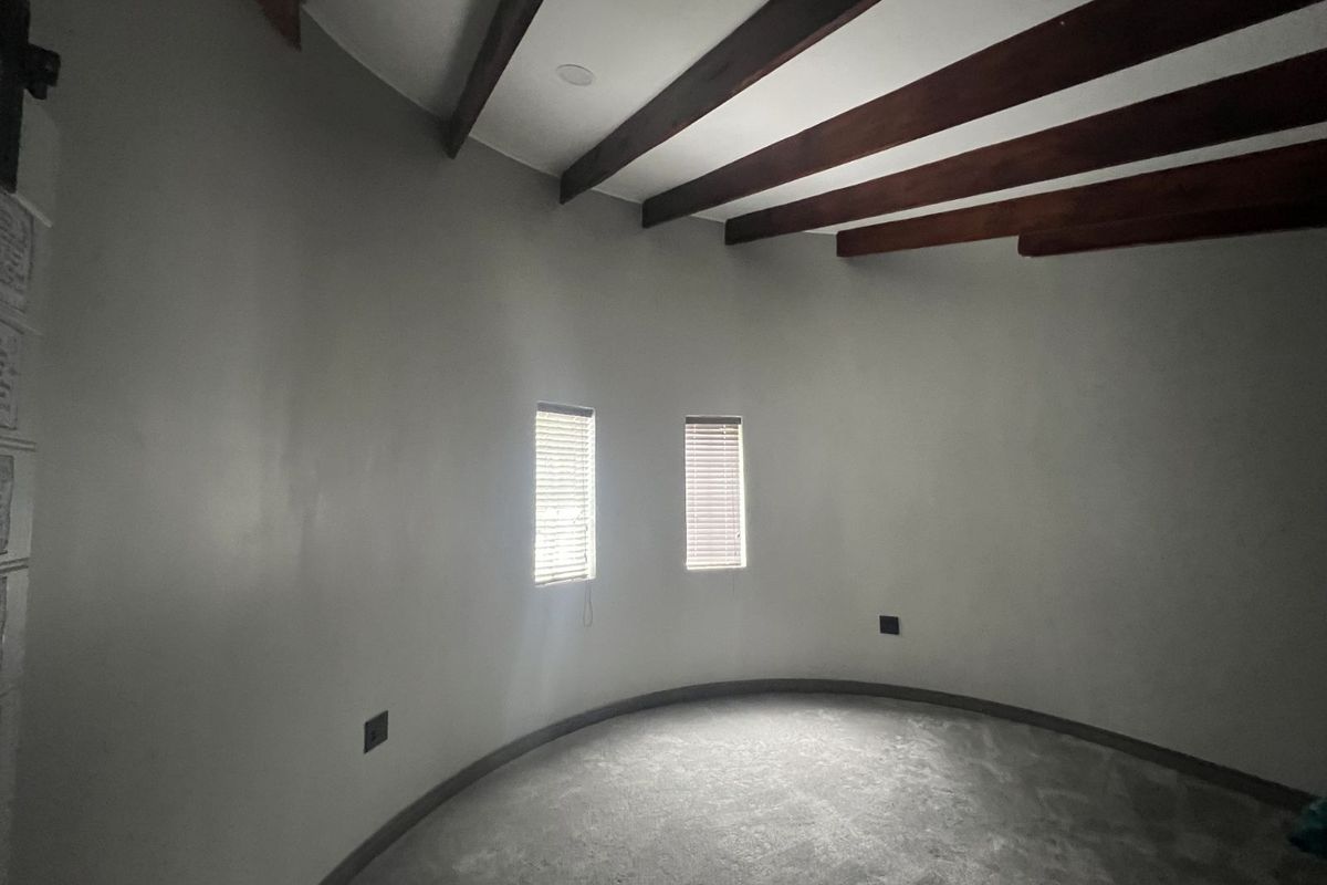 A spacious, empty room featuring a unique curved wall design, exposed timber ceiling beams, and two windows with fitted blinds.