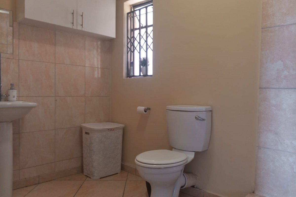 A functional bathroom featuring a pedestal basin, toilet, wall-mounted storage cabinet, and tiled flooring with a security-grilled window.