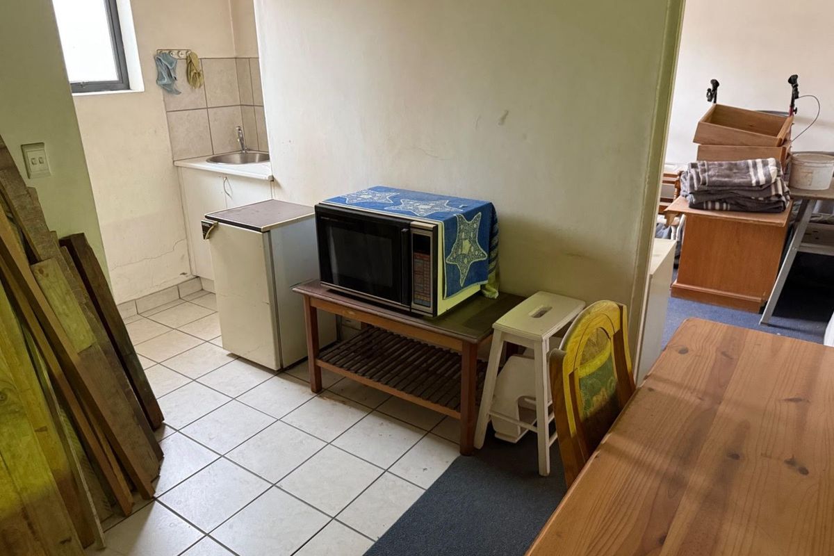 A compact kitchenette area featuring tiled flooring, a small sink unit with tiled splashback, and basic cabinetry in an industrial property setting.