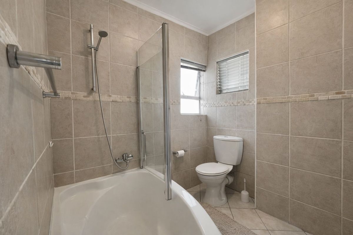 A functional bathroom featuring a bathtub with a glass shower screen, a toilet, and neutral wall and floor tiling.