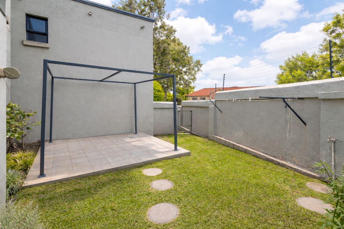 A private rear garden featuring a paved patio area with a metal pergola frame, lawn, and a secure boundary wall with a washing line.