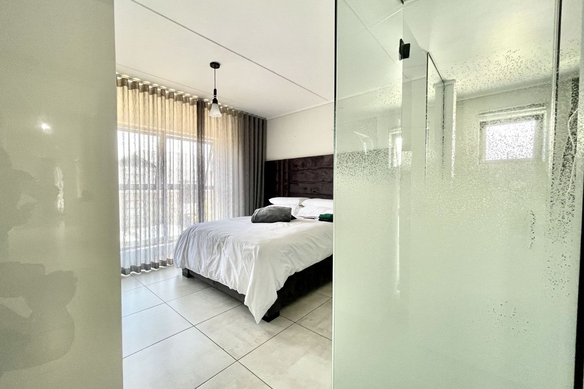 A bright bedroom featuring large sliding glass doors with floor-to-ceiling curtains, tiled flooring, and an adjacent glass-enclosed bathroom area.
