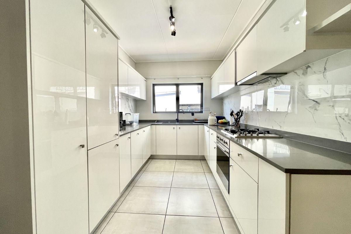 Modern galley-style kitchen featuring high-gloss white cabinetry, stone countertops, marble-effect splashbacks, integrated oven, gas hob, and sleek black hardware.