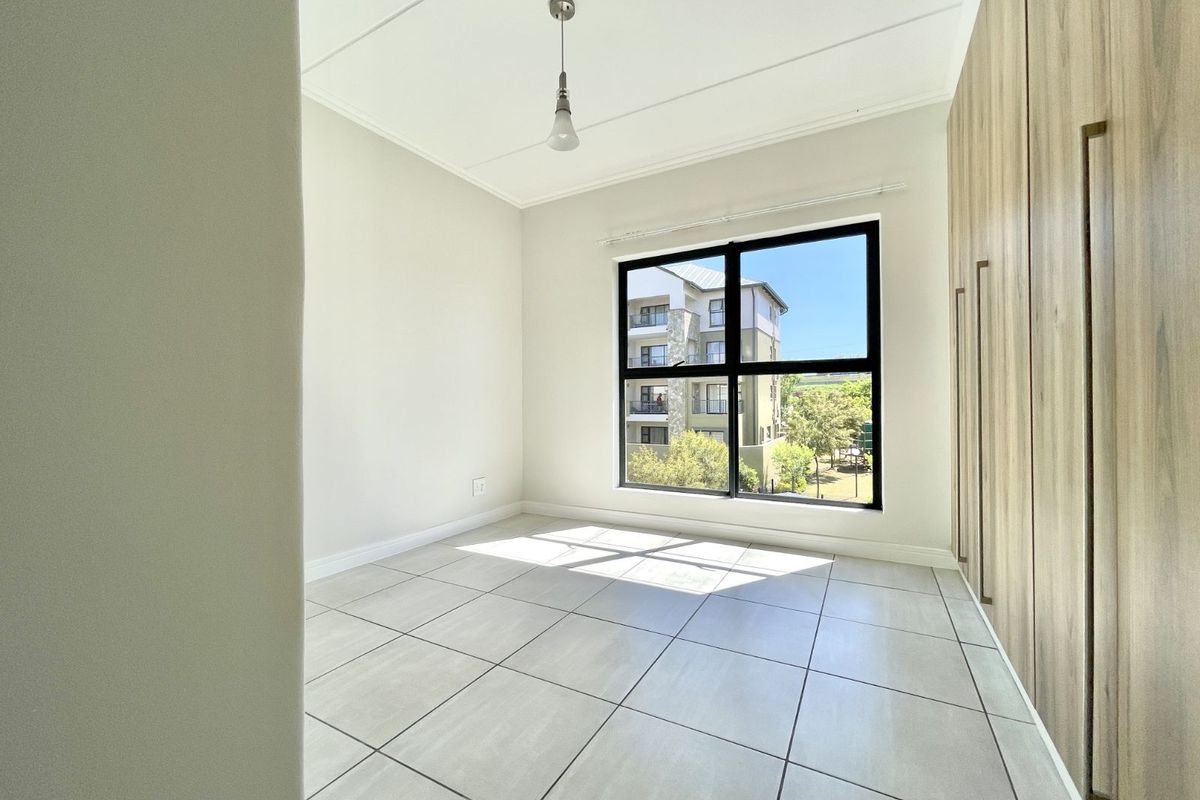 A bright, empty bedroom featuring neutral floor tiles, built-in wooden cupboards, and a large window providing natural light and neighborhood views.