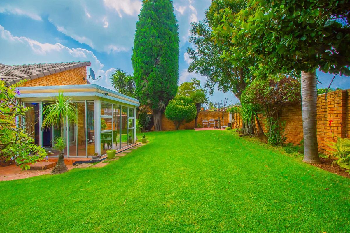 A spacious rear garden featuring a well-maintained lawn, mature trees, brick boundary walls, and an enclosed patio area with sliding glass doors.