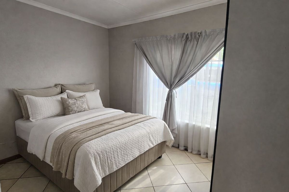 A well-maintained bedroom featuring neutral wall tones, tiled flooring, and large windows with sheer curtains allowing for natural light.