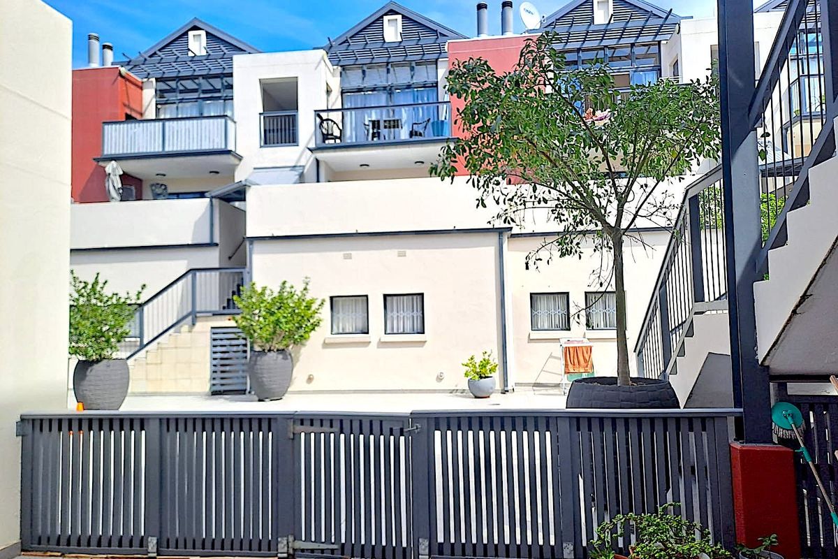 A view of the rear exterior of the apartment complex, featuring multi-level balconies, paved courtyard area, and secure metal fencing.