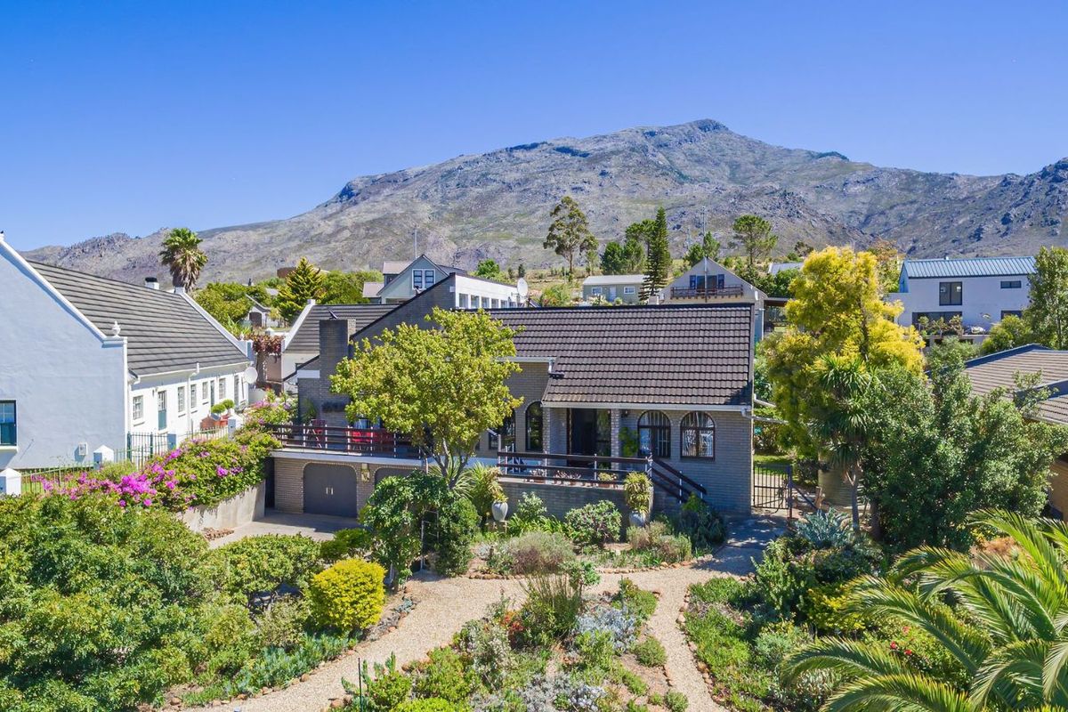 Elevated view of a charming residential property featuring a landscaped garden, paved pathways, and a raised patio area against a scenic mountain backdrop.
