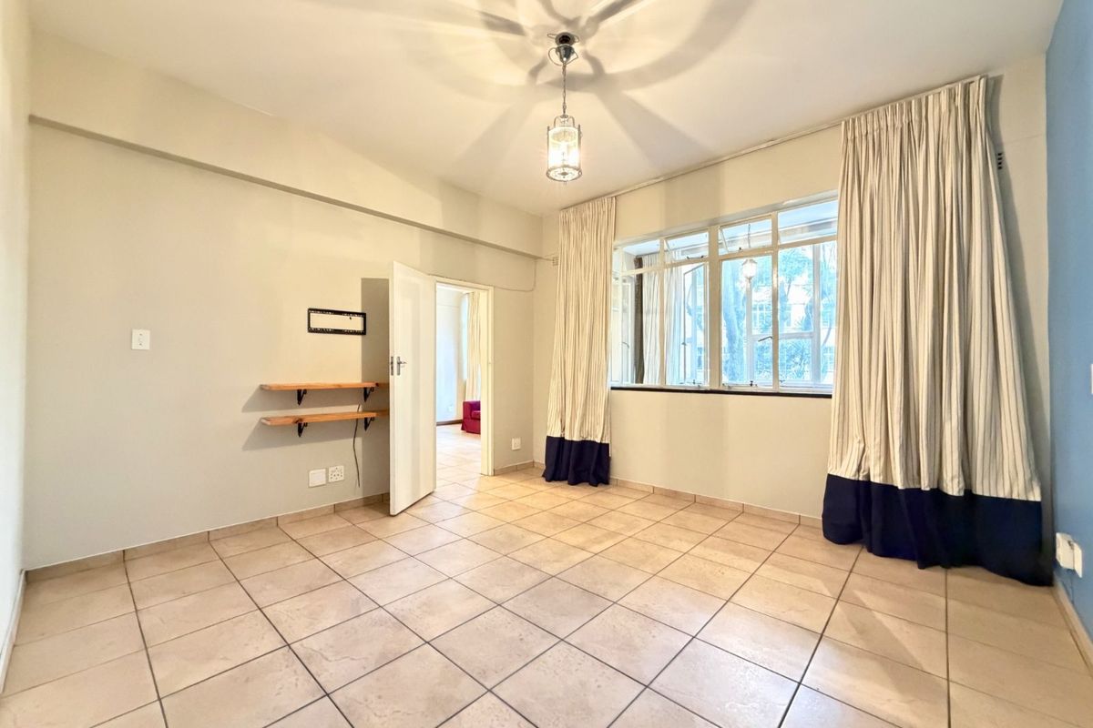 A bright, empty living room featuring neutral tiled flooring, large windows with curtains, and a central pendant light fixture.